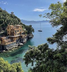 a boat is in the water next to an old house on a cliff near some trees