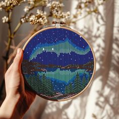 a hand is holding up a cross - stitch pattern with trees and mountains in the background