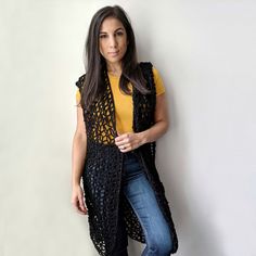 a woman leaning against a white wall wearing a black crochet vest and jeans