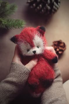 a hand holding a red stuffed animal next to pinecone cones and evergreen needles