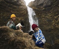 two people sitting on the side of a cliff next to a waterfall with a dog