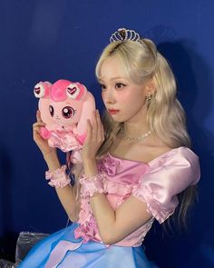 a woman in a pink dress holding up a stuffed animal with big eyes and a tiara on her head