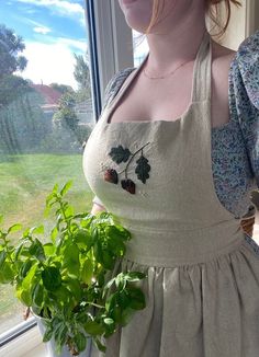 a woman standing in front of a window holding a potted plant and wearing an apron