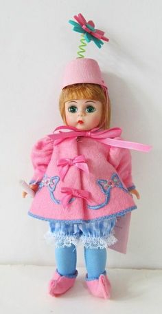 a doll wearing a pink coat and blue pants with a pink hat on it's head