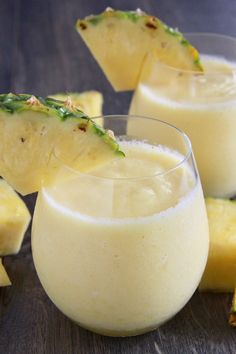 two glasses filled with pineapple drink sitting on top of a wooden table next to sliced pineapples