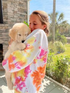 a woman holding a puppy in her arms while wearing a tie - dyed shirt and yellow pants