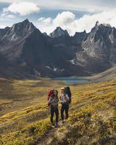 two people with backpacks are walking up a hill towards the mountain lake and mountains