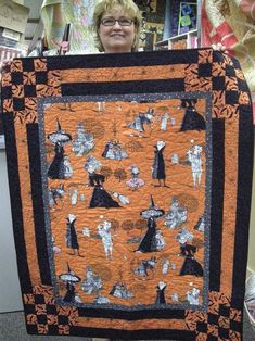 a woman holding up an orange and black quilt with images of witches all over it