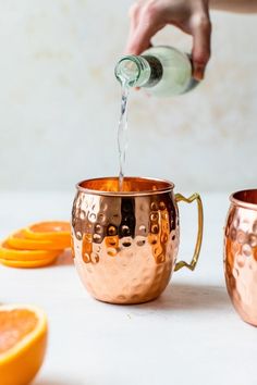 a person pours water into a copper mug with orange slices on the table next to it