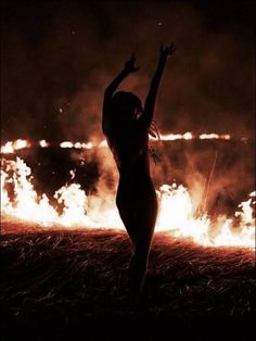 a woman standing in front of a fire with her arms up to the sky,