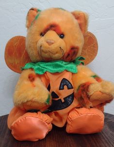 a teddy bear dressed up as a pumpkin sitting on top of a wooden table next to a white wall