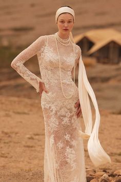 a woman in a white dress and head scarf standing on the beach with her hands on her hips