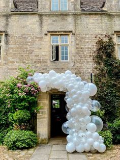 an arch made out of white balloons in front of a brick building with flowers and greenery