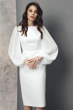 a woman wearing a white dress with long sleeves and an open back, standing in front of a gray wall