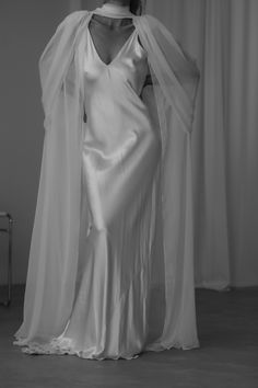 Bride wears a simple slip dress created with silk satin inside a bridal shop. her hair is up in a bridal pony. she styles her wedding dress with a silk chiffon cape. the cape has an ethereal look with draping along the shoulders and an elegant necktie accent. the look is ethereal and luxurious for her modern wedding. Scarf For Wedding Dress, Sheer Top Over Wedding Dress, Sheer Bridal Cape, Wedding Dresses With Shawls, Short Wedding Dress With Cape, Silk Wedding Dress With Veil, Wedding Gown Cape, Wedding Dress With Cape Sleeves, Wedding Dress Cape