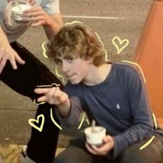 a young man sitting on the ground holding a drink and talking to another person with hearts drawn around him