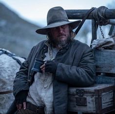 a man wearing a hat and coat sitting on top of a wooden box next to a horse