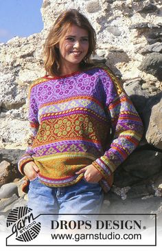 a woman standing in front of some rocks wearing a colorful knitted sweater and jeans