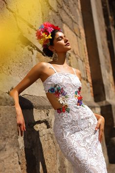 a woman wearing a white dress with flowers in her hair leaning against a stone wall