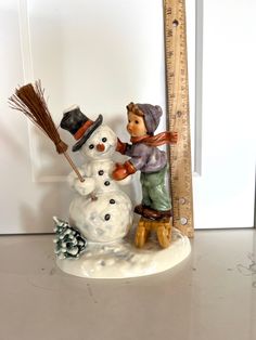 a snowman with a hat and broom next to a tall ruler on a table