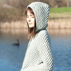 a woman standing in front of a body of water wearing a hooded sweater and looking off into the distance