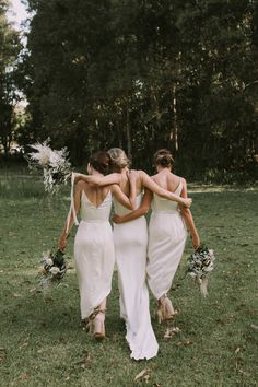 three bridesmaids in white dresses walking through the grass with their backs to each other