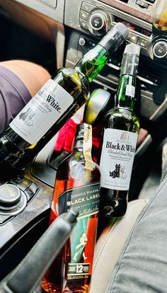 several bottles of alcohol sit in the center console of a car, while someone is holding their hand on the steering wheel