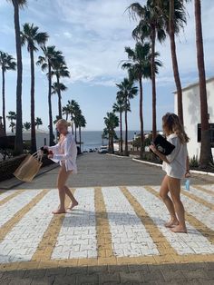 two young women walking down a street next to palm trees and the ocean in the background