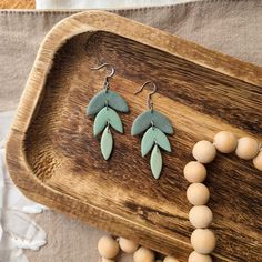 two pairs of earrings with green leaves on them sitting on a wooden tray next to beads