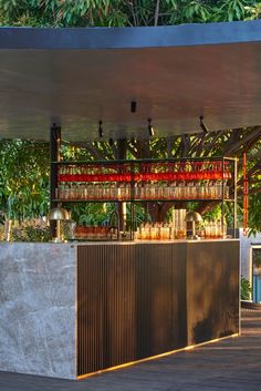 an outdoor bar with lots of bottles on the top and shelves above it in front of some trees