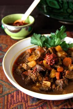 a white bowl filled with meat and vegetables on top of a colorful table cloth next to bowls of soup