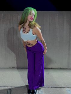 a woman with green hair and tattoos standing in front of a wall wearing purple pants