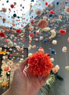 a hand is holding an orange flower in front of a ceiling with flowers hanging from it