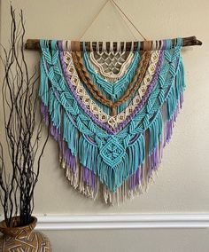 a wall hanging made out of different colored yarns and wood sticks with some plants in front of it