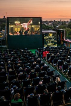 an outdoor movie theater with people sitting in chairs watching the sun set over the water