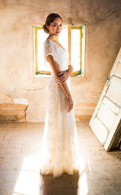 a woman in a white wedding dress standing by a window with sunlight streaming through it