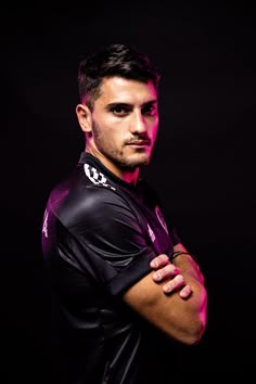 a man with his arms crossed standing in front of a dark background wearing a black shirt