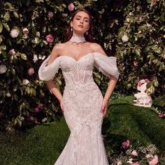 a woman standing in front of flowers wearing a wedding dress with an off the shoulder neckline