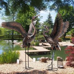 two metal birds standing next to each other on top of a wooden platform in front of a pond