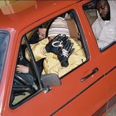 two men sitting in the drivers seat of a red car, one wearing a hat and gloves