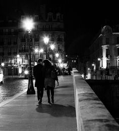 two people walking down a sidewalk at night with street lamps on either side of them