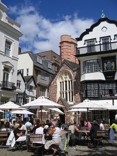 many people are sitting at tables under umbrellas in front of some old buildings on a sunny day