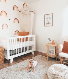 a baby sitting on the floor in front of a white crib with rainbows painted on it