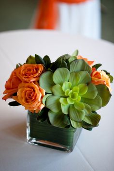 an arrangement of orange roses and green leaves in a square vase on a white table