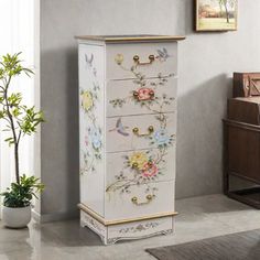 a white cabinet with flowers painted on the front and drawers, next to a potted plant