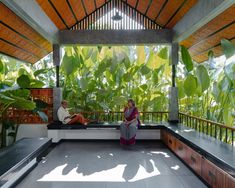 two people sitting on benches in a covered area surrounded by plants and greenery, with sunlight streaming through the roof