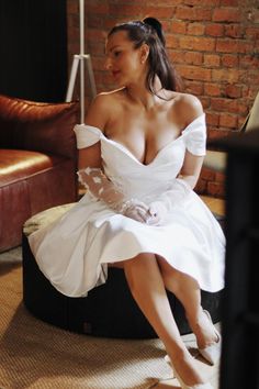 a woman in a white dress sitting on top of a round ottoman next to a brick wall