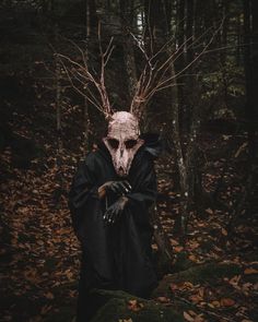 a man in a black robe and mask with horns standing in the woods, holding his hands out