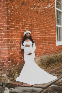 a pregnant woman standing in front of a brick wall wearing a white gown and flower crown