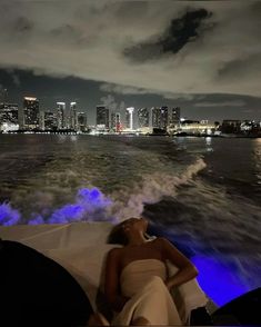 a woman laying on the back of a boat at night with city lights in the background